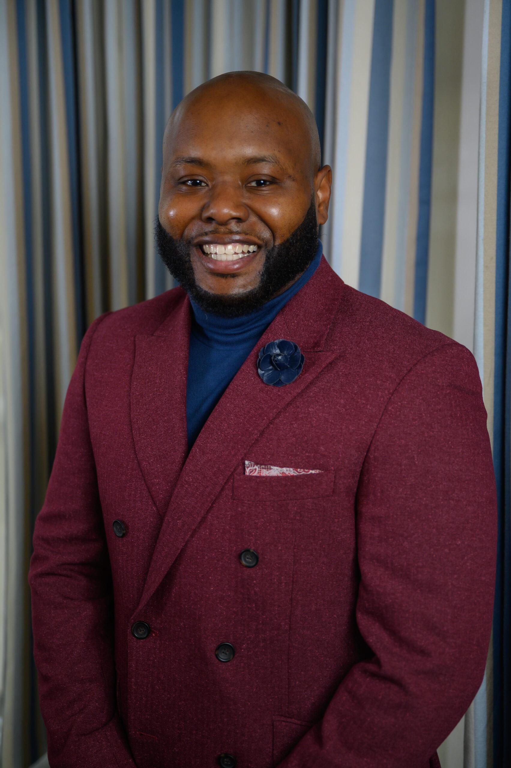 Jarel Jackson stands in front of a gray backdrop. He is wearing a maroon blazer with a navy turtleneck underneath.